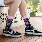 A person sitting outdoors on a wooden bench wearing black Vans sneakers and pink-and-black patterned crew socks. The socks feature colorful graffiti-style graphics and text, with a pink cuff.