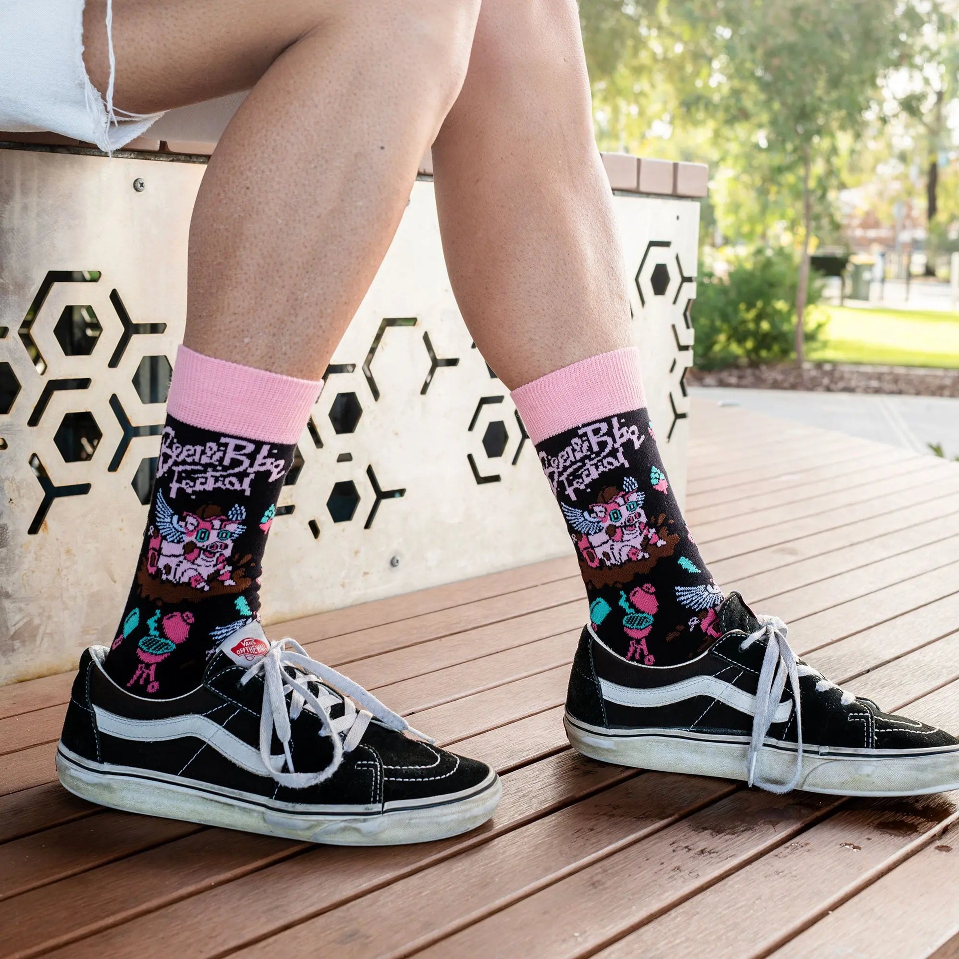 A person sitting outdoors on a wooden bench wearing black Vans sneakers and pink-and-black patterned crew socks. The socks feature colorful graffiti-style graphics and text, with a pink cuff.