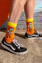 Male model is wearing bright orange and yellow funky novelty socks with cowboy zombies drinking moonshine. 