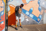 Male model posing next to graffiti wall wearing bright and colourful orange and yellow novelty socks of cowboy zombies drinking moonshine.